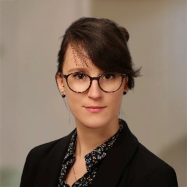 Headshot of Maria standing in front of abstract background smirking at the camera. Wearing glasses with black blazer and paterend blouse