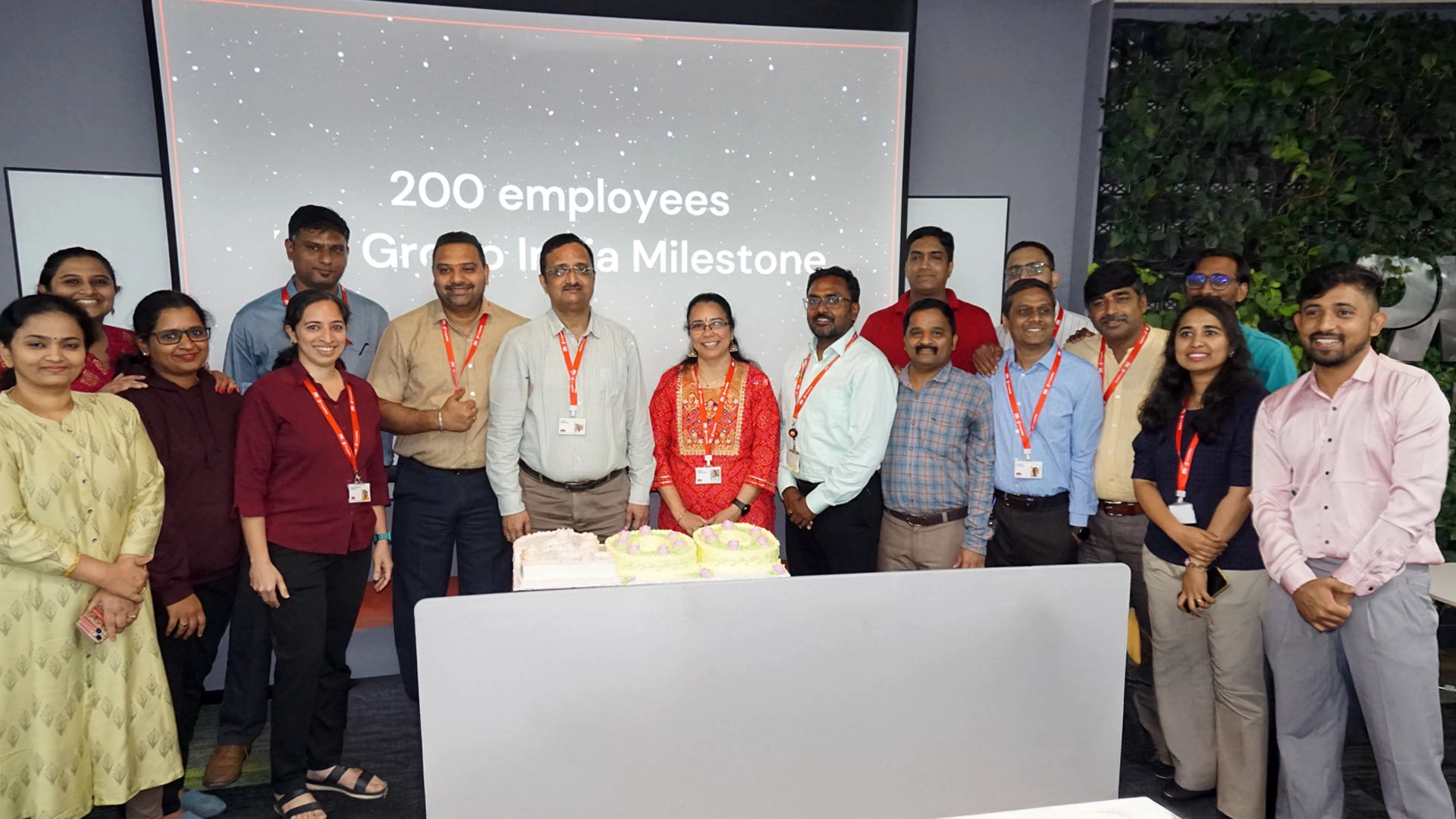 A group of employees standing together in front of a screen that reads ‘200 employees Group Milestone.’ Two cakes are on the table in front of them