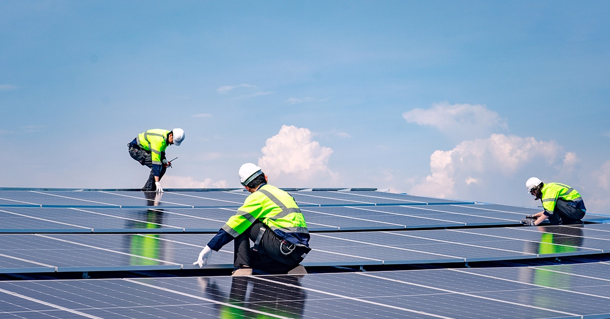 Three people in hi-vis jackets and sfatey gear assessing large solar panels on top of a building