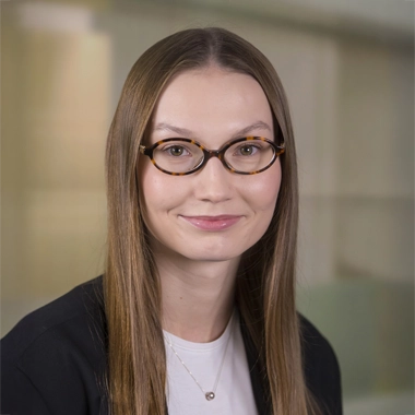 Headshot of Wiktoria standing in front of an abstract background smiling with glasses and black blazer and white top
