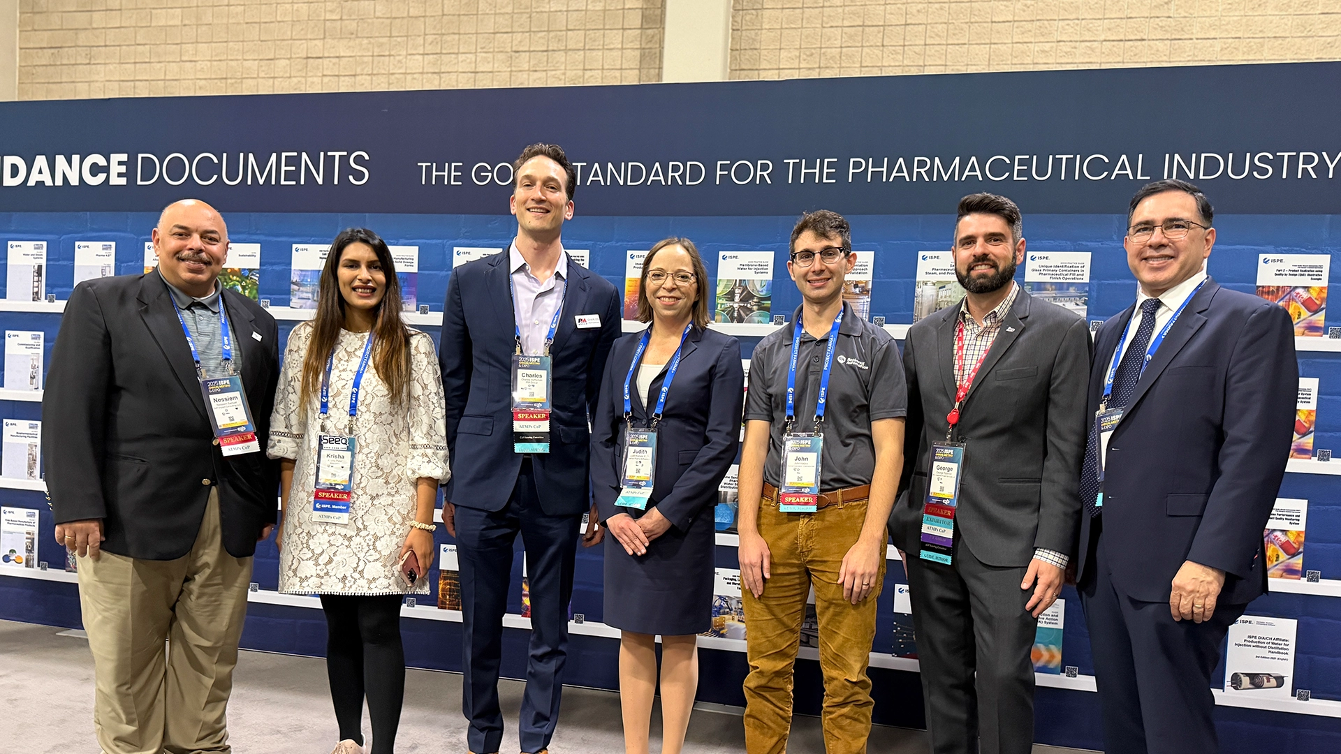 Group of men and women posting for a photo, standing in front of a dark blue wall displaying reports across the wall.
